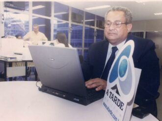 Eliezer Varjão sentado numa mesa da redação do A TARDE, ele veste terno preto com gravata preta. A sua frente uma mesa redonda e um notebook antigo. A sua frente, diante da câmera, uma placa em PVC escrito "A TARDE online".