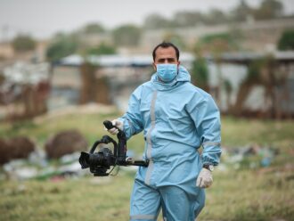 Homem vestido com trajes higiênicos, estilo de hospital na cor azul, usa máscara azul também e carrega uma câmera DSLR em mãos apoiada sob um suporte de cor preta assim como a câmera. Aparentemente encontra-se dentro de um cemitério