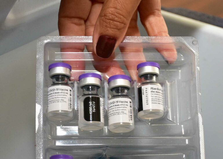 Mãos de uma técnica de enfermagem de unhas pequenas e pintadas de vinho seguram uma embalagem plástica com 4 imunizantes em pote de vidro e tampa roxa